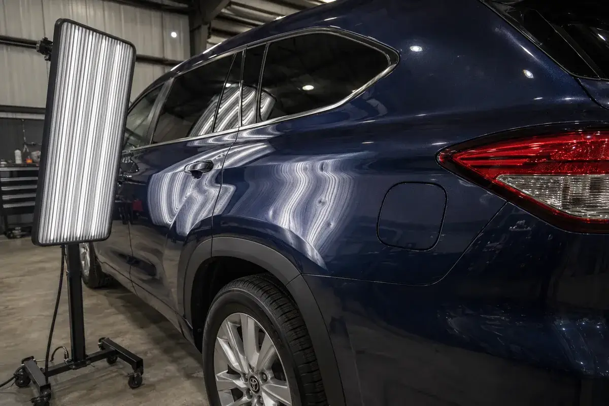 A blue Toyota Highlander quarter panel under a striped reflection board during paintless dent repair at Podium Body Works in Round Rock, Texas