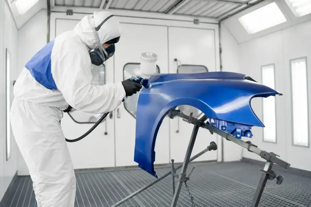 A painter in a spray suit refinishing a blue fender in the paint booth at Podium Body Works