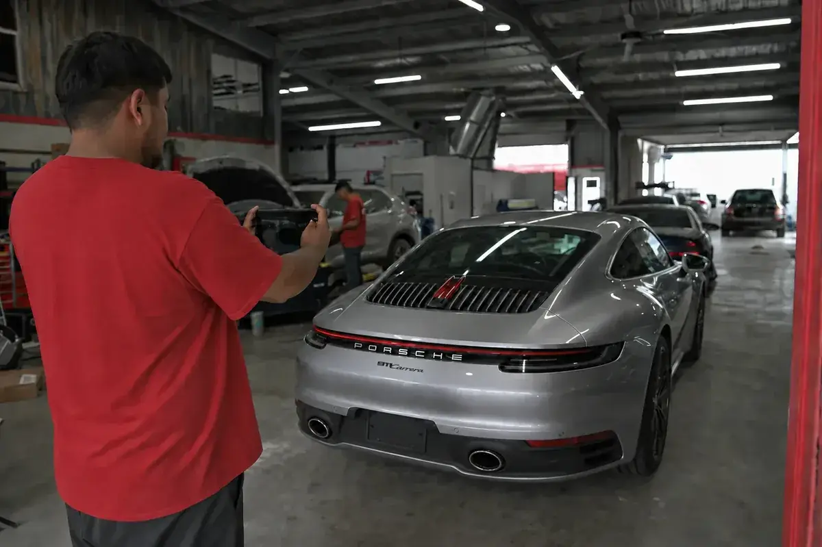 A staff member photographing a silver Porsche 911 at intake for an estimate at Podium Body Works in Round Rock, Texas