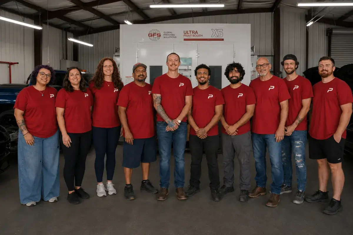 The Podium Body Works team in red shirts standing in front of the paint booth at the shop in Round Rock, Texas